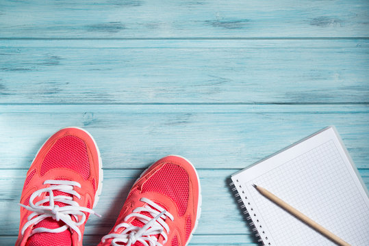 Fitness concept, pink sneakers and notebook with pencil on wooden background, top view