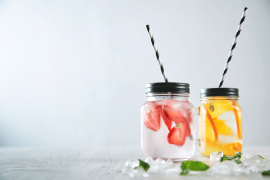 Close up two fresh homemade lemonades made from sparkling water, ice, strawberry and orange. Melted crashed ice and mint leaves around, striped drinking straw inside rustic jars. Cold beverages