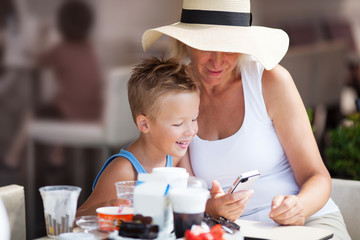 Smiling Mature Woman and Young Boy On Vacation Using Cell Phone Together on Outdoor Restaurant Patio