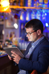 Businessman wearing glasses sitting at a table reading a tablet in a brightly lit colorful cocktail bar, close up side view