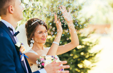 elegant stylish groom with his happy gorgeous blonde bride have fun with soap bubble on the background of trees outdoors in park