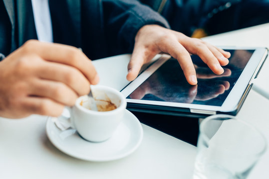 Close up on the hands of young businessman stirring with a tablespoon a cup of coffee and using tablet, tapping screen - technology, work, break concept
