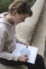 Young woman drawing a purple mandala pattern on a sketchpad with a pencil outdoors with the sea...