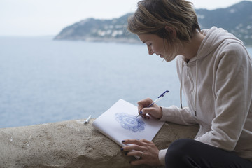 Young woman drawing a purple mandala pattern on a sketchpad with a pencil outdoors with the sea...