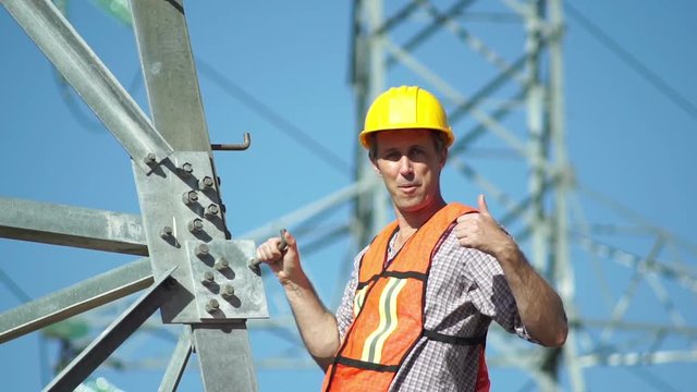 Close up shot of a male technician in a safety vest and hard hat standing on the side of a high tension electrical tower visually inspects the situation and gives the viewer a thumbs up gesture.