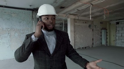 Young african american manager of the construction company goes through the premises of repaired building, talking on the smartphone. This is handsome young man in a business suit and construction