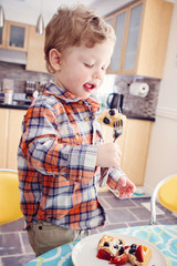 Portrait of one happy kid boy eating breakfast waffles with fruits with fork in sunny kitchen early morning showing his tongue