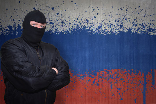dangerous man in a mask standing near a wall with painted national flag of russia