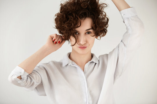 Portrait of attractive Caucasian hipster girl posing against white conrete wall with pounting lips. Student female with short curly hairstyle having fun while dancing to some pleasant music indoor