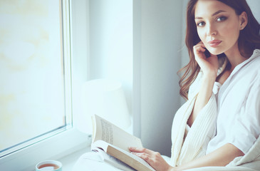 Young woman at home sitting near window relaxing in her living room reading book and drinking coffee or tea