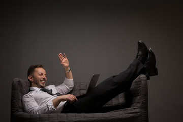 Happy businessman lying on comfortable sofa with laptop computer. Smiling man waving to photographer in studio. Studio shot.