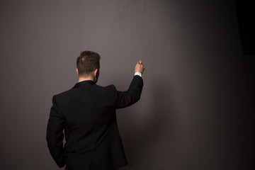 Back view of young businessman writing something on wall in studio. Handsome man in black suit posing for photographer with one hand in pocket.