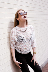 Fototapeta premium Beautiful blond young hipster girl with red lipstick and bright makeup on a sunny day outdoors. Smiling sexy woman dressed in a black leisure jacket posing near old buildings in France, Italy, Spain.