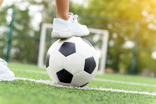 Feet of a boy wearing white sneakers stepping on a soccer ball in the middle of the football field.