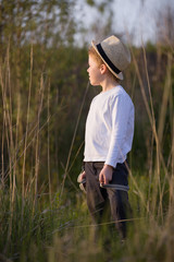 Adorable kid boy in straw hat standing on a summer meadow and looking into distance. Sunset in the park. Outdoors. Vacations.
