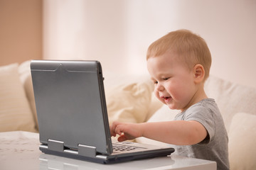 Cute little toddler boy sitting on the sofa with his laptop and looking at the window. Learning with computer. Early development. Child and computer.