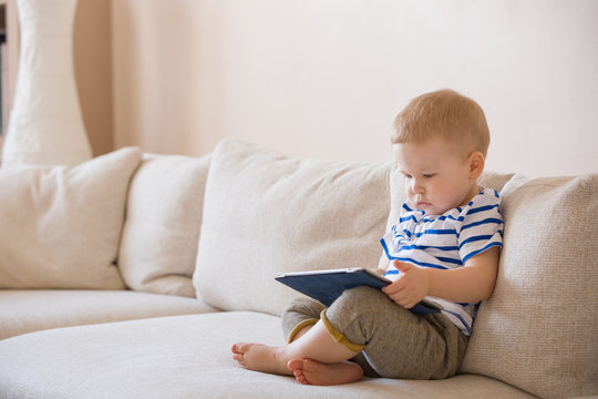 Adorable blond toddler boy laying on the sofa and playing with tablet pc at home, indoors. Child with tablet computer.
