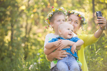 Portrait of adorable kids with their young mother taking selfie in the sunny park. Happy family in flower wreaths making photo with smartphone outdoors on a summer day. Boys and mom smiling.