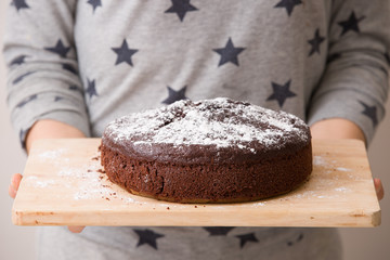 Homemade dark chocolate birthday cake with cream, chocolate chips on its top. Cake with white sugar powder in woman's hands ready for party and celebrations. Grey with dark blue stars as a background.