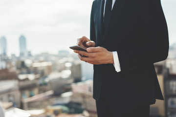 Cropped image of professional businessman using smartphone outside, confident emloyer in black suit standing on the roof of modern business centre and typing text message on his cellphone