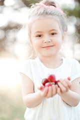 Fototapeta premium Smiling baby girl 3-4 year old holding strawberries outdoors closeup. Looking at camera. Childhood. Healthy eating.