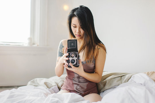 Young woman sitting on bed, taking photograph with vintage camera