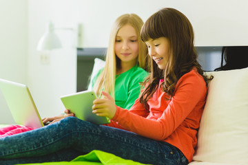 Two beautiful little sisters sitting on bed and play with a Tablet or laptop