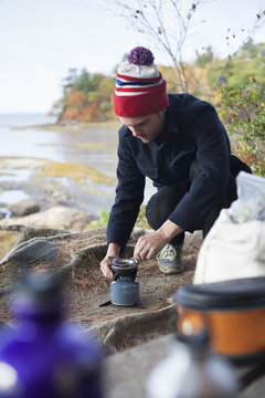 Male hiker igniting camping stove on riverbank, Rangeley, Maine, USA