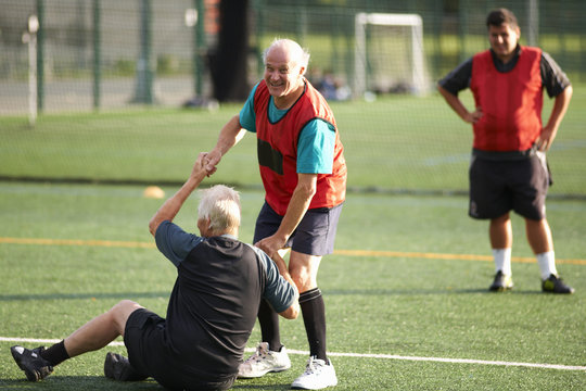 Senior man helping friend stand up on football pitch