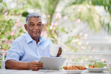 Portrait of smiling aged man with tablet enjoying morning coffee