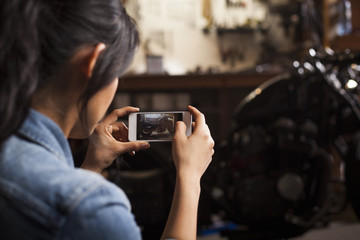 Female mechanic in workshop, taking photograph of motorcycle using smartphone