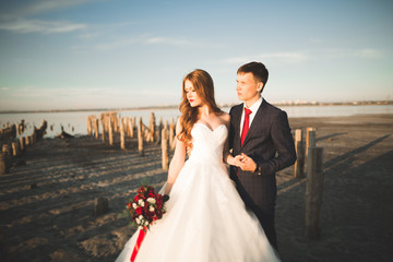 Wedding couple, groom, bride with bouquet posing near sea on sunset