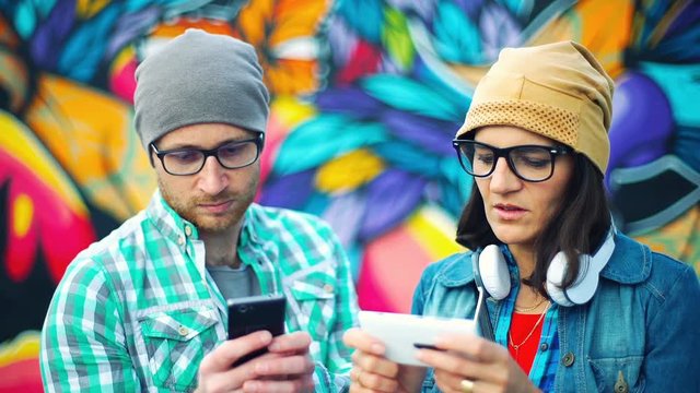 Pair pf hipsters standing next to each other and using smartphones

