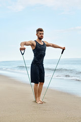 Sports. Sporty Handsome Male Doing Expander Exercise During Outdoor Workout Near Sea. Fit Athletic Man With Muscular Body Exercising, Training At Ocean Sand Beach. Fitness, Healthy Lifestyle Concept