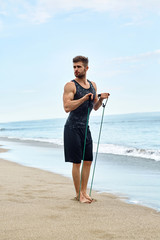 Exercise. Handsome Fit Athletic Man With Muscular Body Doing Stretching Expander Exercises On Sand. Sportsman Exercising At Beach. Sporty Male Training During Outdoor Workout. Sports, Fitness Concept