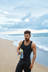 Portrait Of Healthy Athletic Man With Fit Body Holding Bottle Of Refreshing Water, Resting After Workout Or Running At Beach. Thirsty Male With A Drink After Outdoor Training. Sports, Fitness Concept