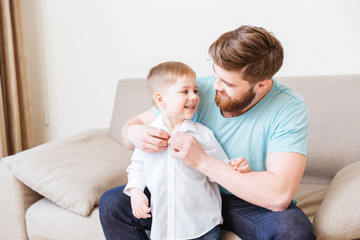 Happy father sitting on sofa and dressing his little son - Powered by Adobe