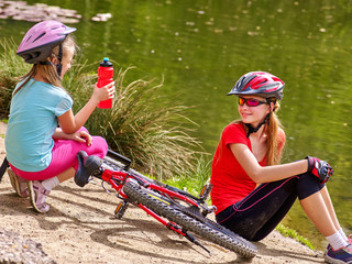 Obraz premium Bikes cycling family. Happy mother and daughter rides bicycle. Family have rest near water in park. Kid drinking bottle water. Bicycles are on coast. 