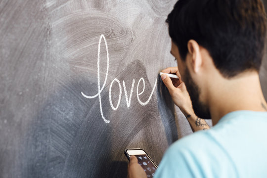 Young man, writing the word love on blackboard, holding smartphone, rear view
