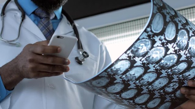 Male african american doctor radiologist viewing mri brain scan film and using smartphone for internet surfing
