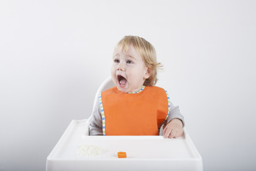 blonde caucasian baby seventeen month age orange bib grey sweater eating rice carrot on white high-chair laughing and smiling

