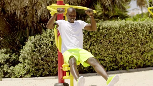 Seated black man exercises on metal equipment painted bright yellow and red near hedges and tropical trees
