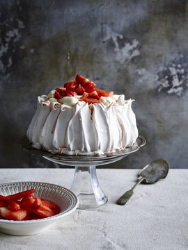 Strawberry covered pavlova on glass cake stand with cake server and bowl of strawberries