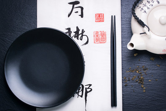 Asia Food Composition With Chinese Chopsticks, Teapot, Dry Green Tea And Empty Plate On A Dark Stone Background With Chinese Hieroglyph, Top View, Toned, With Free Space For Text