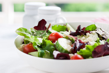 Summer season salad with salad leaves, tomatoes, cucumbers, Italian herbs and cheese in a bowl on a table, closeup, selective focus 