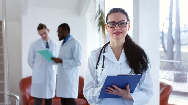 Friendly doctor in glasses. Female caucasian doctor in glasses writing in clipboard, looking at camera and siling, with two male doctors at the background