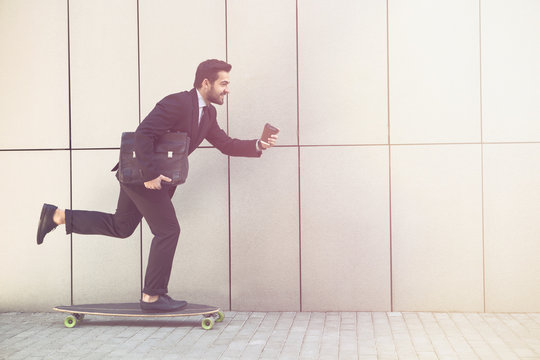Toned picture of businessman on longboard and with cup of coffee going to office. Freelance man in black business suit hurrying.