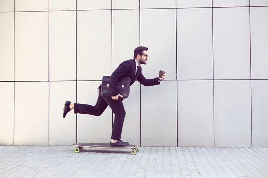 Toned image of businessman in black businesssuit on longboard walking with cup of coffee in his hand. Freelance man hurrying to his office.