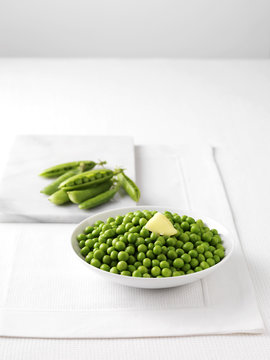Raw pea pods on marble cutting board and bowl of boiled peas with butter