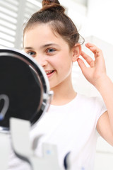 Aparat słuchowy dla dziecka. Wesoła dziewczynka zakłada aparat słuchowy przeglądając się w lusterku - obrazy, fototapety, plakaty